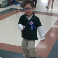 little boy with Down syndrome wearing a blue shirt and brown pants and blue glasses with a pin that says Birthday boy