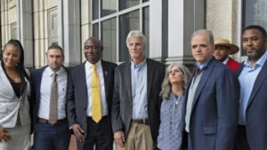 A professional photograph showing Matthew Dietz, Aaron Karger and Benjamin Crump standing in front of a modern glass-fronted building with a group of colleagues. He is wearing a dark suit and tie, smiling, and positioned in the center among his associates.