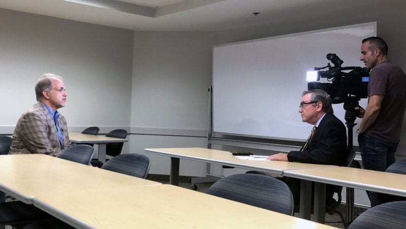 Prof. DIetz in a brown jacket and blue shirt being interviewed with a cameraman and a reporter who is wearing glasses and a black suit.
