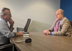 Professor Dietz in a tan checkered suit and red tie, being interviewed by a white man in a checkered shirt with a microphone on the table
