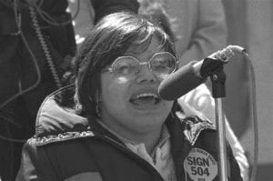 black and white photo of a woman with glasses wearing a warm coat with a pin that says Sign 504, she is speaking into a microphone