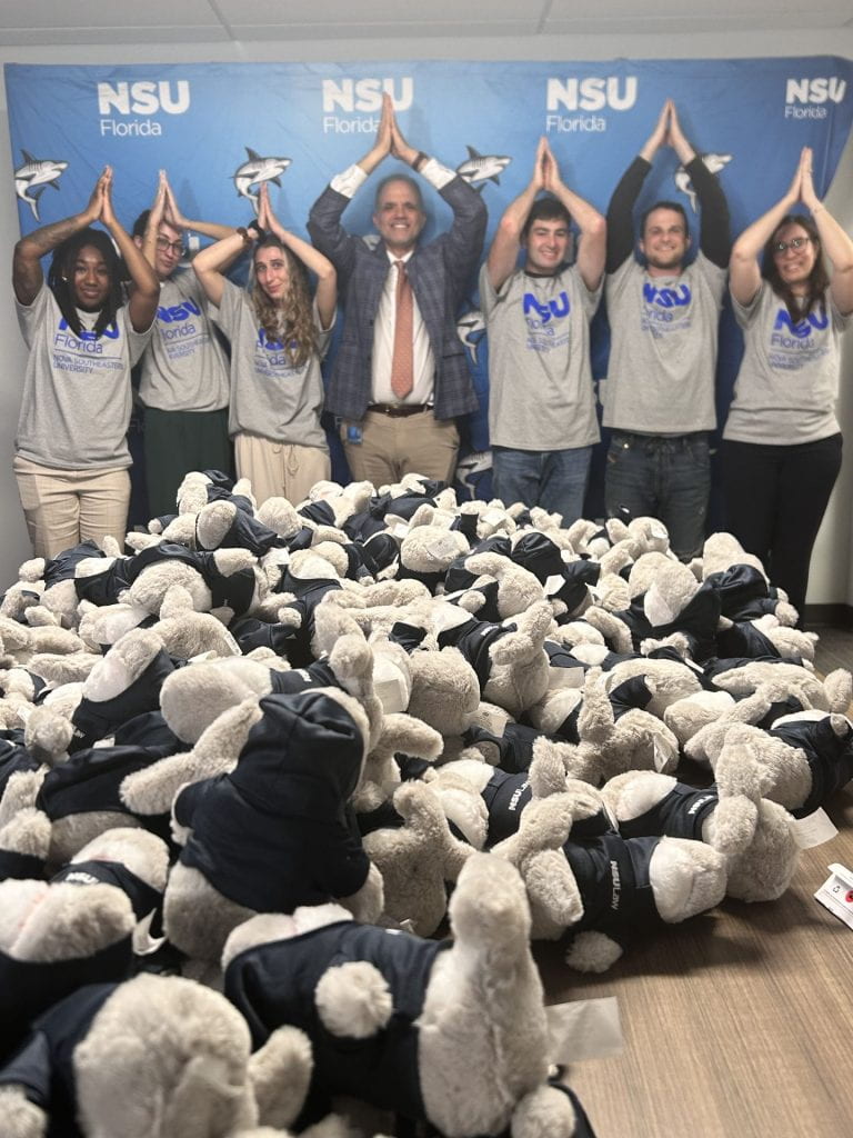 six students with NSU t-shirts and Professor Dietz - all with their arms in the air forming a fin, woith hundreds of small stuffed sharks in the foreground.